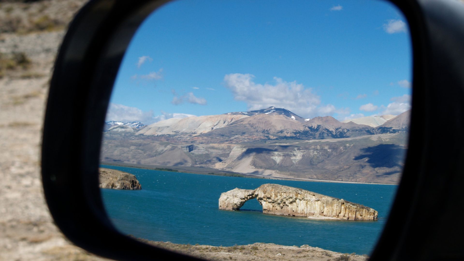 Une photo d'un rétroviseur de voiture montrant le lac Posadas et sa célèbre arche rocheuse reflétée dans le miroir