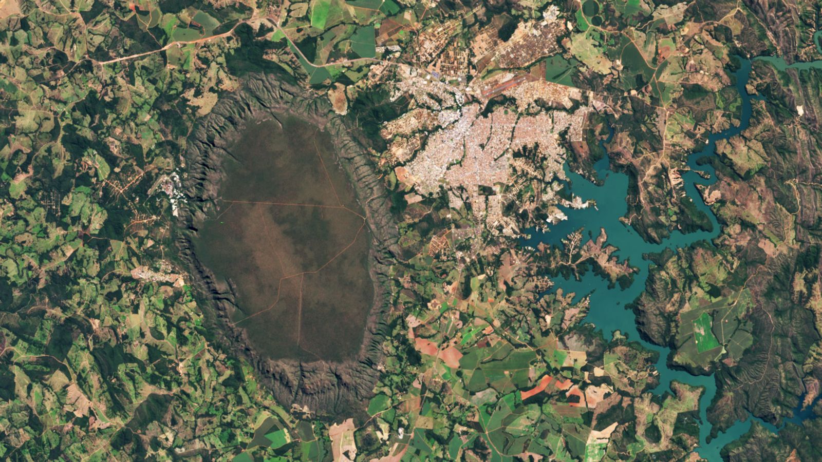 A satellite photo of a giant dark dome next to a town and an artificial lake, all surrounded by patchwork fields