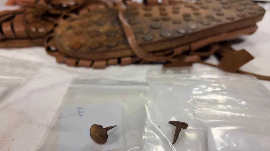 two rusted metal hobnails sit on a table in the foreground with reconstructed Roman hobnail sandals in the background