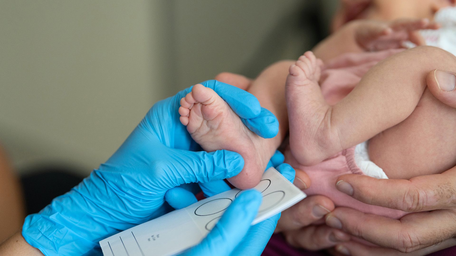 Une main gantée de bleu mesure le pied d'un bébé.