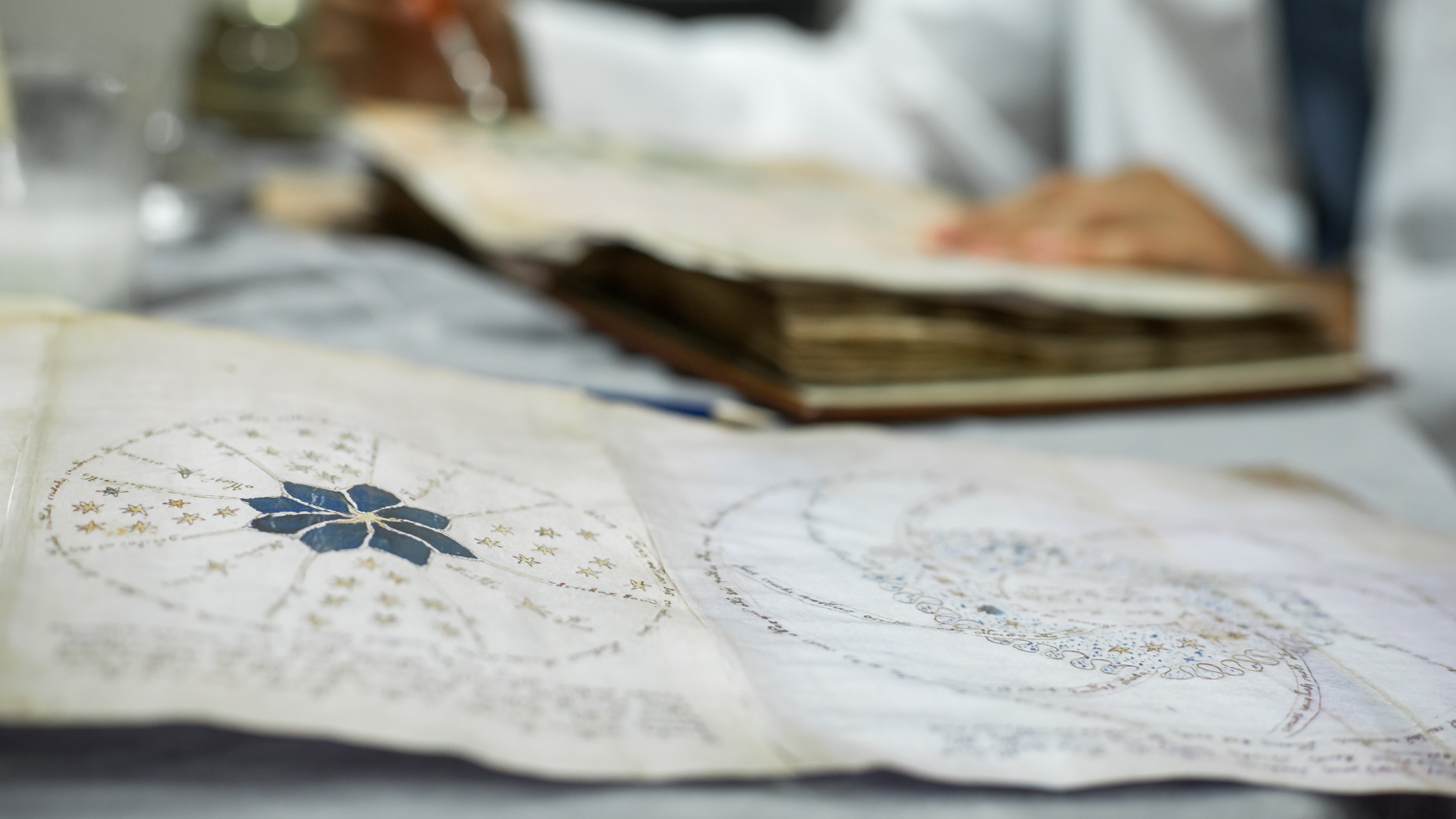 a person works on a manuscript in the background while the foreground has a page of parchment from the Voynich manuscript