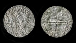A close up of the back and front of a silver coin; one side shows a man's profile and the other shows a cross.