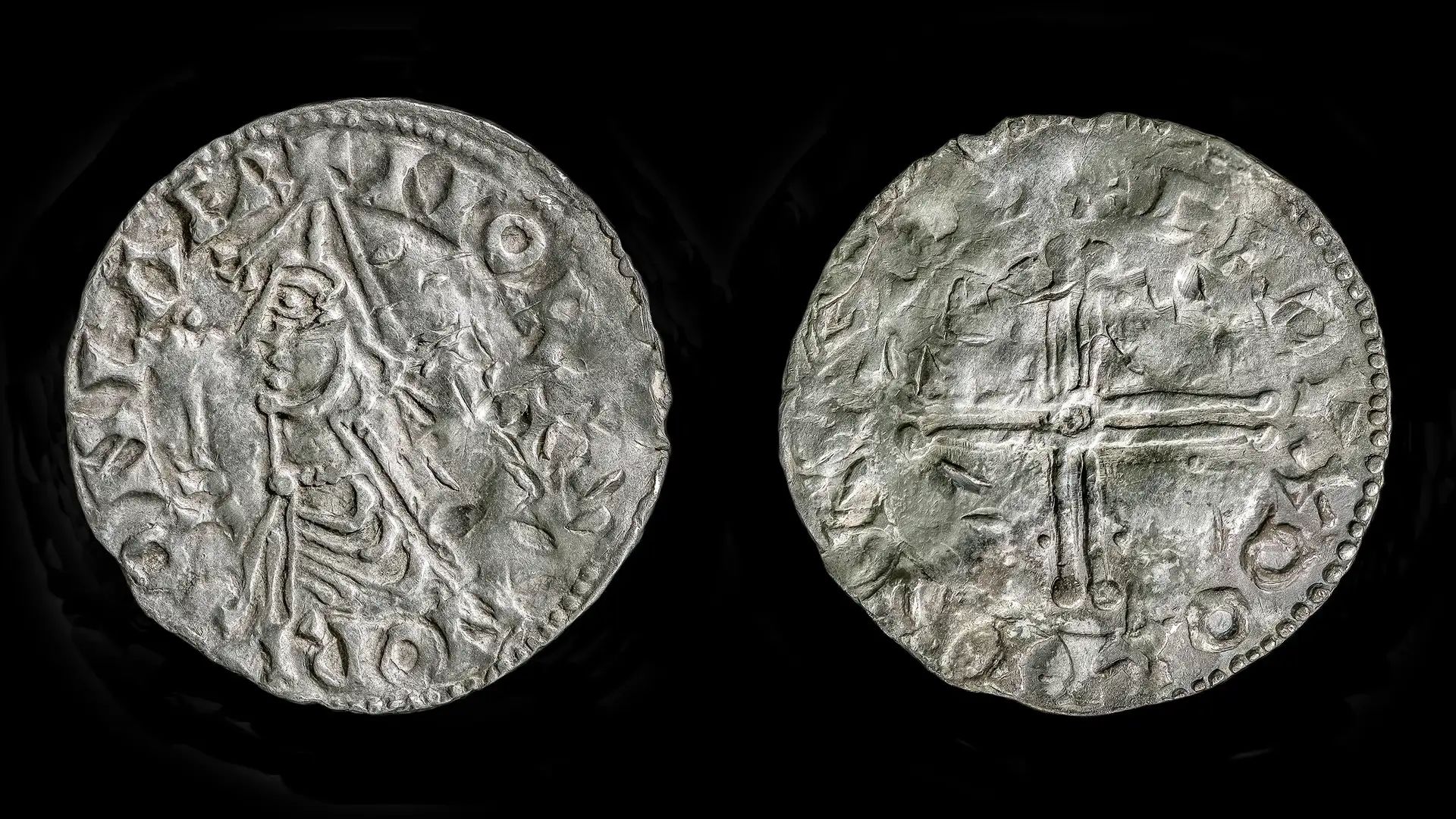 A close up of the back and front of a silver coin; one side shows a man's profile and the other shows a cross.