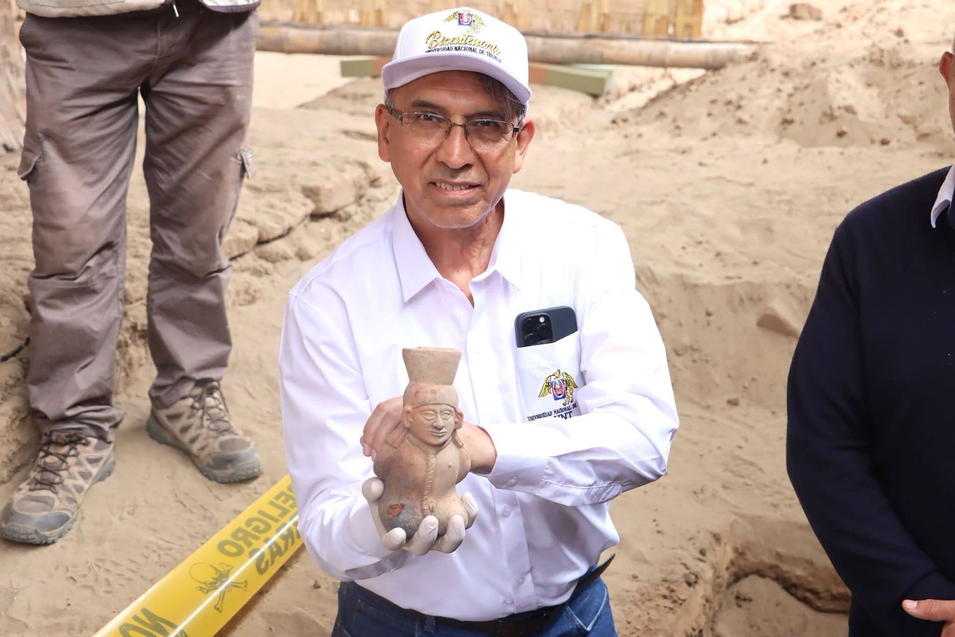 Un homme vêtu d’une jolie chemise et d’une casquette de baseball tient un morceau de poterie Moche sur un chantier de fouilles.