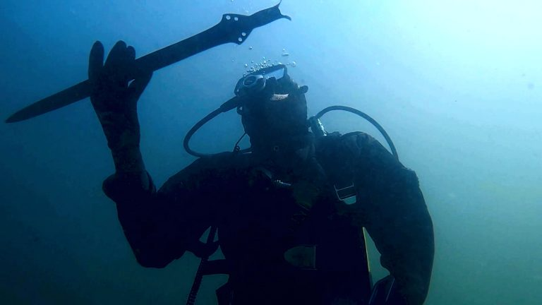 an underwater photograph shows a person wearing a dive suit holding a Bronze Age sword